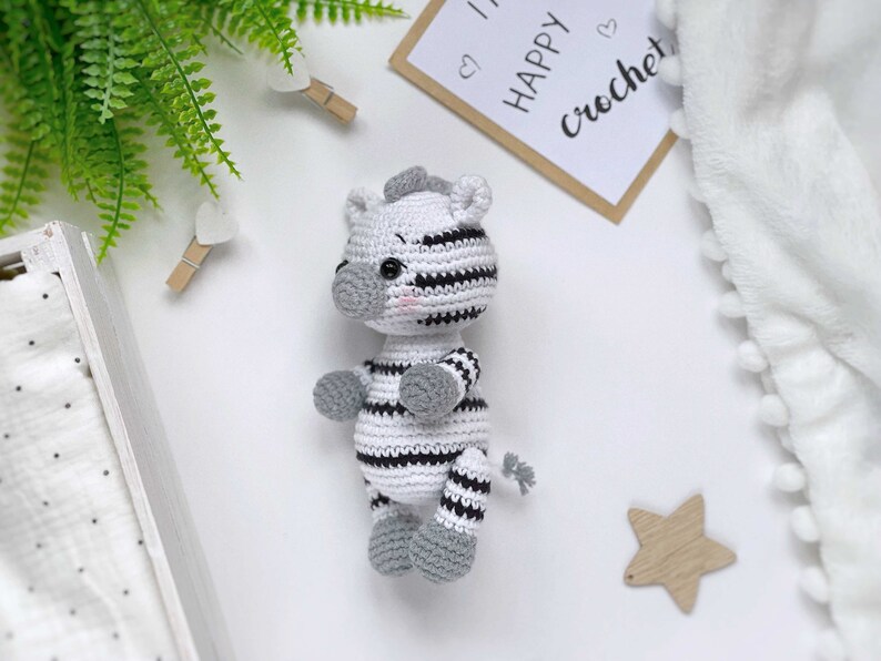 May include: A small, handmade crochet zebra toy featuring a white body with black horizontal stripes, a grey snout, grey ears, grey feet, and a small grey tail. The zebra has black safety eyes and subtle pink blush details on its cheeks.