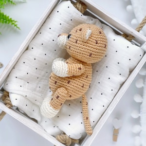 Op de afbeelding: Een gehaakt knuffeldier in de vorm van een luipaard met beige garen, donkerbruine vlekken en wit garen op de pootjes en de snuit. Het speelgoed ligt op een witte stoffen doek met kleine zwarte stippen in een witte houten kist met touwgrepen.