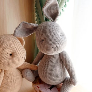 Op de afbeelding: Twee gehaakte knuffels, een grijs konijn en een beige beer. Het konijn zit op een kleine, ronde doos met een bloemenpatroon. De beer staat naast het konijn.
