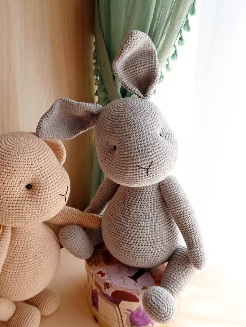 Op de afbeelding: Twee gehaakte knuffels, een grijs konijn en een beige beer. Het konijn zit op een kleine, ronde doos met een bloemenpatroon. De beer staat naast het konijn.
