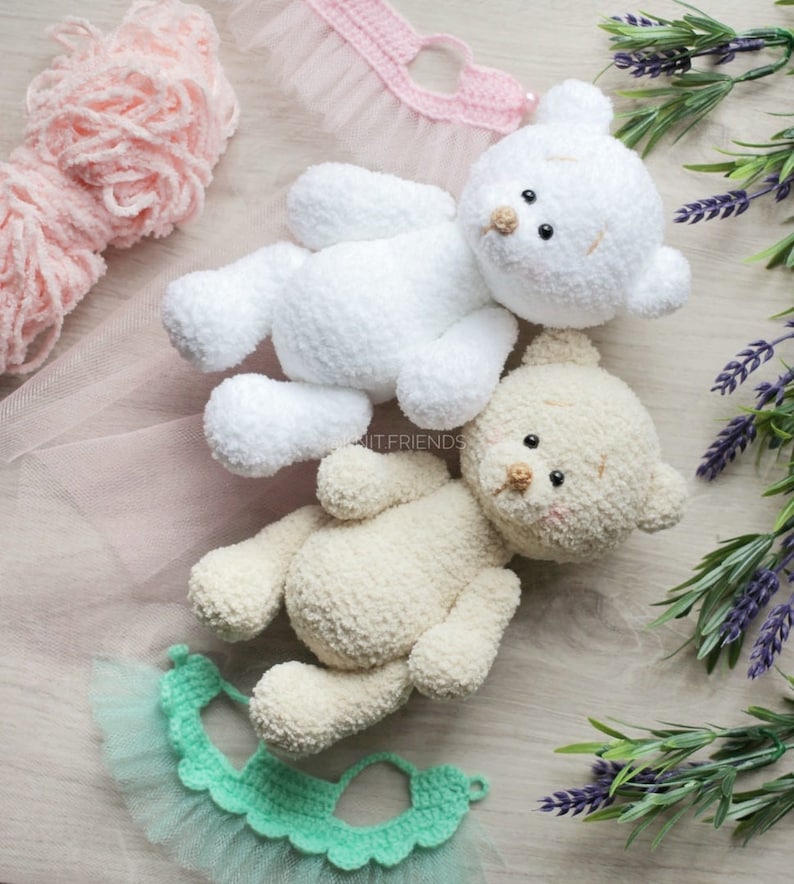 May include: Two crocheted teddy bears, one white and one beige, with black eyes and brown noses. The bears are lying on a light wood surface with a pink and green crocheted tutu.