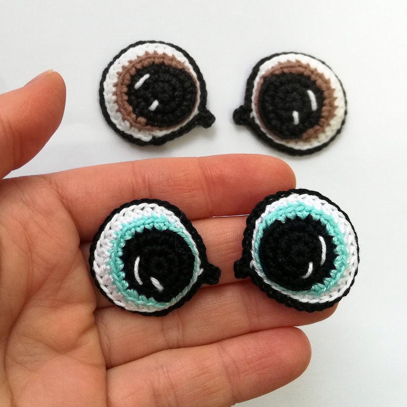 May include: Four crocheted eyes with black pupils and white and colored accents. Two eyes have brown and white accents, and two eyes have turquoise and white accents.