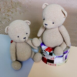 Peut inclure: Deux ours en peluche beige crochet&eacute;s. L'un est assis sur une bo&icirc;te ronde d&eacute;corative avec des motifs floraux et textuels. L'autre est assis sur une surface bleu clair. Les deux ours ont des yeux noirs et des d&eacute;tails cousus.