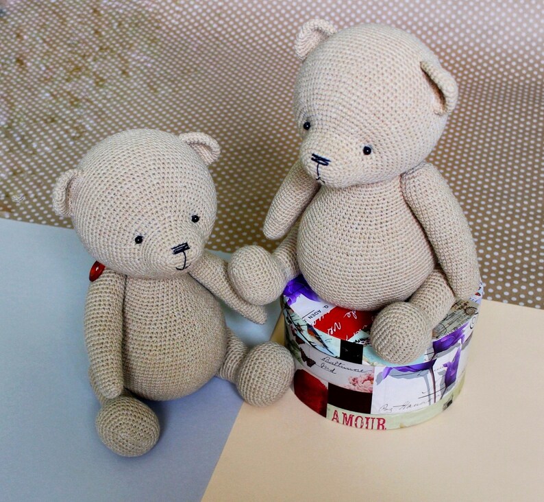 Peut inclure: Deux ours en peluche beige crochet&eacute;s. L'un est assis sur une bo&icirc;te ronde d&eacute;corative avec des motifs floraux et textuels. L'autre est assis sur une surface bleu clair. Les deux ours ont des yeux noirs et des d&eacute;tails cousus.
