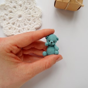 Könnte beinhalten: Ein kleiner, türkisfarbener Häkelteddybär mit schwarzen Augen und einer gestickten Nase. Der Bär sitzt in der Handfläche.