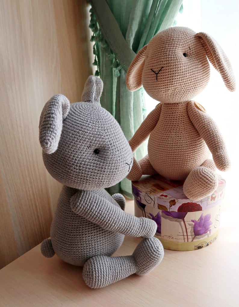 Op de afbeelding: Twee gehaakte knuffels van konijnen, een grijs en een lichtbruin, zittend op een doos met bloemenprint. Het grijze konijn zit op de vloer en het bruine konijn zit op de doos.