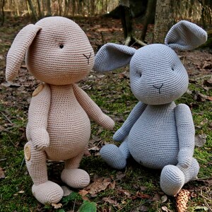 Op de afbeelding: Twee gehaakte konijnenknuffels, een in lichtbruin en een in grijs, zittend op een bed van bladeren en mos.
