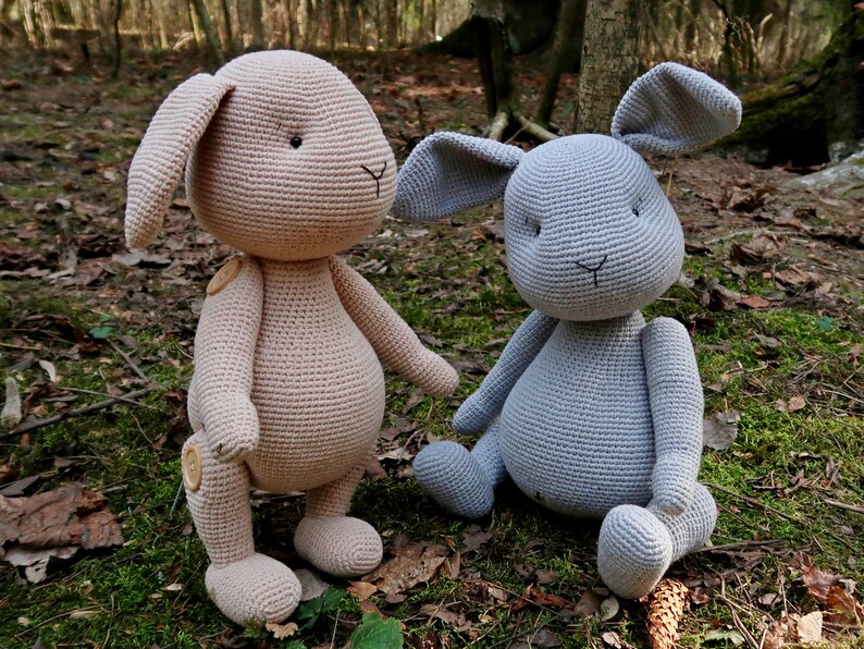 Op de afbeelding: Twee gehaakte konijnenknuffels, een in lichtbruin en een in grijs, zittend op een bed van bladeren en mos.