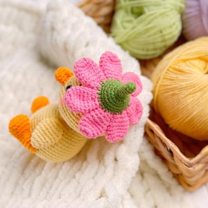 May include: A small, yellow, crocheted duckling toy featuring orange feet and a beak, with a large, pink, crocheted flower attached to its head. The flower has a green center and stem-like base, and the duckling has small black eyes and pink cheeks.