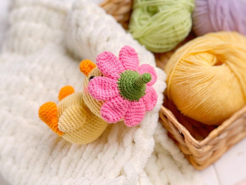 May include: A small, yellow, crocheted duckling toy featuring orange feet and a beak, with a large, pink, crocheted flower attached to its head. The flower has a green center and stem-like base, and the duckling has small black eyes and pink cheeks.