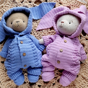 May include: Two crocheted stuffed toys wearing matching outfits. The toy on the left is wearing a blue outfit with a hood and the toy on the right is wearing a purple outfit with a hood.