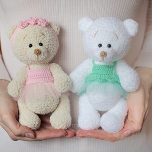 May include: Two crocheted teddy bears wearing tutus. One bear is cream colored with a pink tutu and a pink flower crown. The other bear is white with a green tutu.
