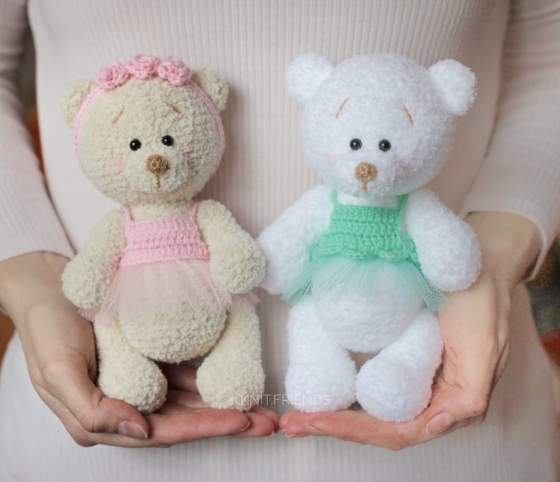 May include: Two crocheted teddy bears wearing tutus. One bear is cream colored with a pink tutu and a pink flower crown. The other bear is white with a green tutu.