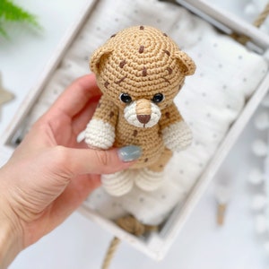 Op de afbeelding: Een klein gehaakt luipaardspeeltje met een beige lichaam, donkerbruine vlekken, een witte snuit en witte pootjes. Het knuffeldier heeft ronde oortjes, een donkerbruin geborduurde neus en zwarte plastic veiligheidsoogjes van garen.