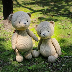 Peut inclure: Deux ours en peluche au crochet beiges faits &agrave; la main sont assis sur de l'herbe verte. Le plus grand ours a un bouton sur sa poitrine. Les ours ont des yeux et des nez cousus noirs. L'arri&egrave;re-plan est une vue floue d'une for&ecirc;t.