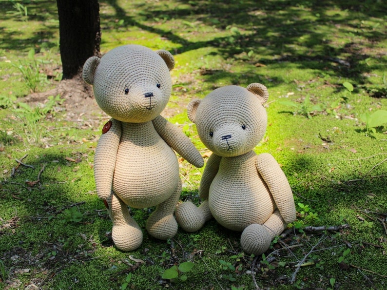 Peut inclure: Deux ours en peluche au crochet beiges faits &agrave; la main sont assis sur de l'herbe verte. Le plus grand ours a un bouton sur sa poitrine. Les ours ont des yeux et des nez cousus noirs. L'arri&egrave;re-plan est une vue floue d'une for&ecirc;t.
