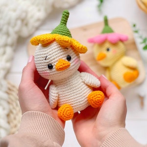May include: A cream-colored crocheted duckling toy featuring a yellow flower-shaped hat with a green stem top, orange feet, and a small orange beak. The toy has black bead eyes and pink blush details on its cheeks, crafted with visible yarn stitches.