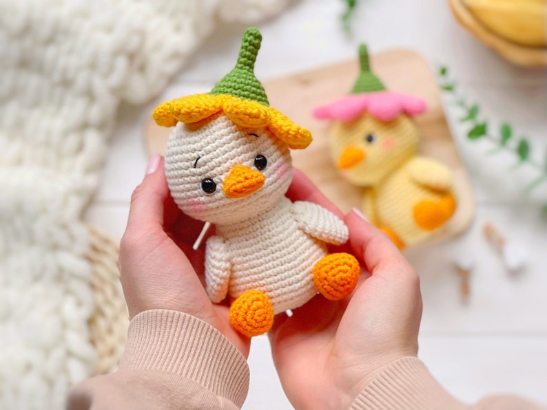 May include: A cream-colored crocheted duckling toy featuring a yellow flower-shaped hat with a green stem top, orange feet, and a small orange beak. The toy has black bead eyes and pink blush details on its cheeks, crafted with visible yarn stitches.