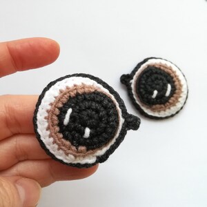 May include: Two crocheted eyes with brown, black, and white yarn. The eyes are round and have a simple face with a small mouth.