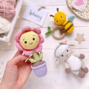 May include: A crocheted flower pot with a pink and yellow flower, a crocheted bee with black and yellow stripes, and a crocheted lamb with a pink bow. The toys are on a white surface with the word "Spring" written on a card.