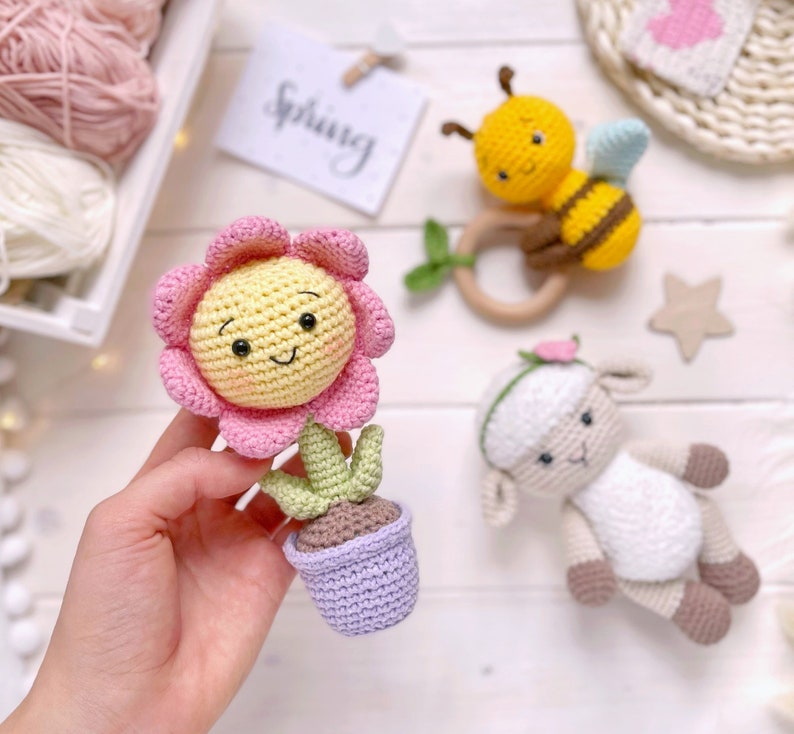 May include: A crocheted flower pot with a pink and yellow flower, a crocheted bee with black and yellow stripes, and a crocheted lamb with a pink bow. The toys are on a white surface with the word "Spring" written on a card.