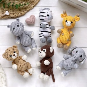 Op de afbeelding: Een verzameling van zes gehaakte knuffeldieren, waaronder een grijs nijlpaard, een zwart-wit gestreepte zebra, een gele giraf met bruine vlekken, een beige luipaard met bruine vlekken, een bruine aap en een grijze olifant, met een klein roze hartje.