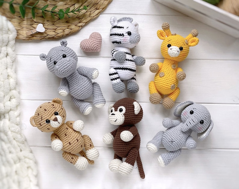 Op de afbeelding: Een verzameling van zes gehaakte knuffeldieren, waaronder een grijs nijlpaard, een zwart-wit gestreepte zebra, een gele giraf met bruine vlekken, een beige luipaard met bruine vlekken, een bruine aap en een grijze olifant, met een klein roze hartje.