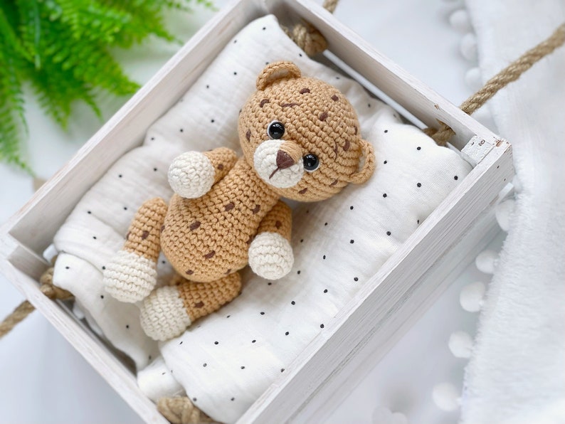 Op de afbeelding: Een gehaakt luipaard-speeltje van beige garen met donkerbruine vlekken en cr&egrave;mekleurige pootjes. Het luipaard heeft zwarte veiligheidsoogjes en een bruin geborduurde neus. Het ligt op een witte mousseline doek met zwarte stippen in een houten kist.