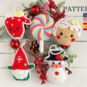Può includere: Cinque ornamenti natalizi all'uncinetto appesi a un ramo con bacche rosse. Gli ornamenti sono un guanto, una canna di zucchero, un omino di pan di zenzero, un gnomo e un pupazzo di neve. Gli ornamenti sono tutti bianchi con dettagli colorati. Il testo "PATTERN" è in alto nell'immagine, con le bandiere americana e spagnola a destra.