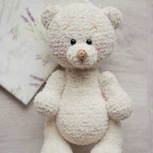 May include: A white crocheted teddy bear with black eyes and a brown nose. The bear is sitting on a light wood surface.