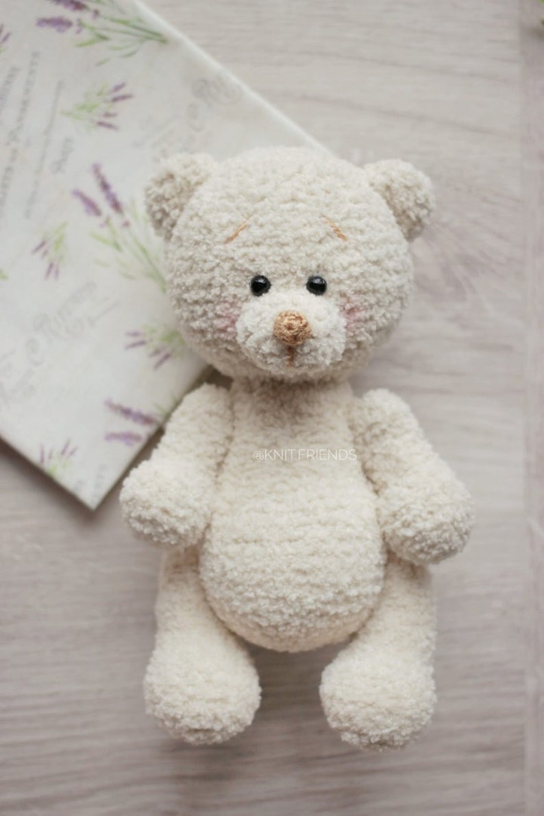 May include: A white crocheted teddy bear with black eyes and a brown nose. The bear is sitting on a light wood surface.