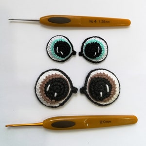 May include: Two sets of crocheted eyes, one set with a teal iris and one set with a brown iris. The eyes are displayed next to two crochet hooks, one labeled "No. 4 1.25 mm" and the other labeled "2.0 mm".
