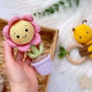 May include: A crocheted flower pot toy with a pink flower and green stem. The pot is purple and the flower has a yellow center with a smiling face.