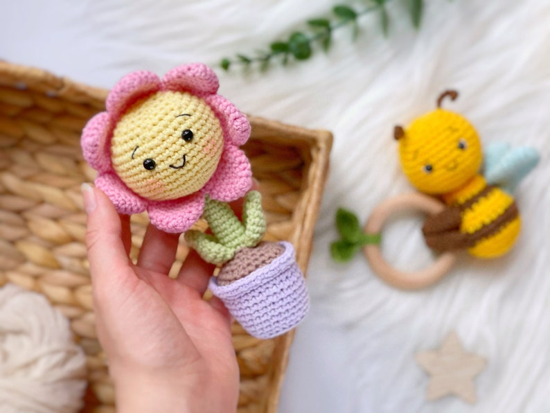 May include: A crocheted flower pot toy with a pink flower and green stem. The pot is purple and the flower has a yellow center with a smiling face.