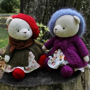 May include: Two handmade crochet teddy bears sit on a tree stump. One bear wears a red beret, green coat, and floral dress. The other bear is dressed in a purple coat and a blue hat, with a floral dress. Both bears have brown noses and are sitting outside.
