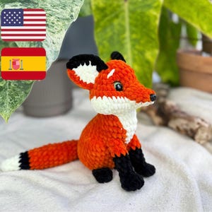 Pode incluir: Um brinquedo de pelúcia de raposa de crochê laranja e branco com olhos pretos e pés pretos. A raposa está sentada em uma superfície branca.