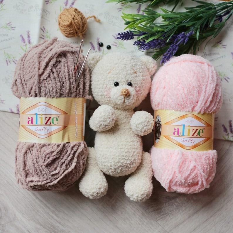 May include: Two skeins of yarn, one pink and one brown, with a crocheted teddy bear in the center. The yarn is labeled "Alize Softy" and is for crocheting.