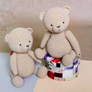 Peut inclure: Deux ours en peluche au crochet beige. Un ours est assis sur une bo&icirc;te d&eacute;corative avec le mot "AMOUR" visible. Les ours ont des yeux noirs et des bouches cousues. Un ours a un bouton rouge sur la poitrine.