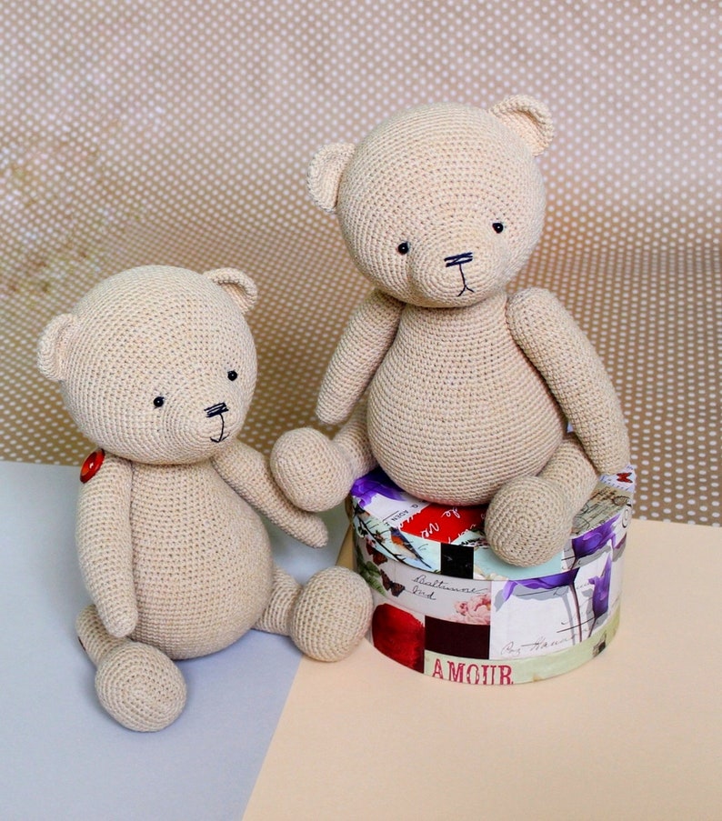 Peut inclure: Deux ours en peluche au crochet beige. Un ours est assis sur une bo&icirc;te d&eacute;corative avec le mot "AMOUR" visible. Les ours ont des yeux noirs et des bouches cousues. Un ours a un bouton rouge sur la poitrine.
