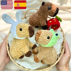 Op de afbeelding: Een rieten mand bevat drie gehaakte capibara's, één met konijnenoren, één met een kikker masker en één met een aardbei. De capibara's zijn bruin en beige, met zwarte ogen. De aardbei is rood.