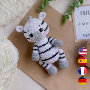 May include: A small, handmade crochet zebra toy featuring a white body with black stripes, gray hooves, and a gray snout. The plush animal has large black safety eyes, embroidered eyelashes, and soft pink blush on its cheeks. It is crafted with yarn.