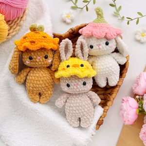 Pode incluir: Três animais de pelúcia de crochê em uma cesta de vime. Os animais são um coelho marrom usando um chapéu laranja, um coelho branco usando um chapéu de flor rosa e um coelho cinza usando um chapéu de pintinho amarelo.
