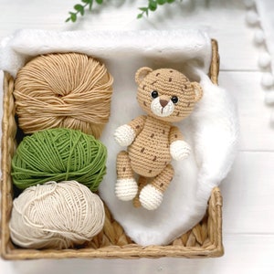 Op de afbeelding: Een gehaakt luipaardspeeltje met bruine vlekken en witte pootjes ligt in een gevlochten mand naast drie bollen garen in beige, olijfgroen en cr&egrave;me. Het luipaard heeft donkere ogen en een bruin geborduurde neus op een zachte witte stof.