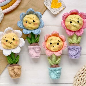 May include: Four crocheted flower pot decorations with smiling faces. The flowers are yellow with white, blue, and pink petals. The pots are pink, brown, purple, and blue.