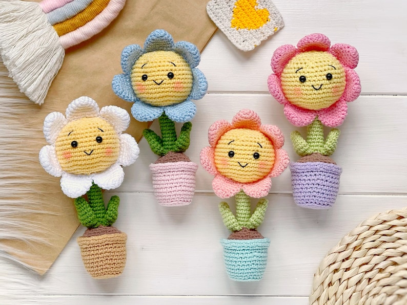 May include: Four crocheted flower pot decorations with smiling faces. The flowers are yellow with white, blue, and pink petals. The pots are pink, brown, purple, and blue.