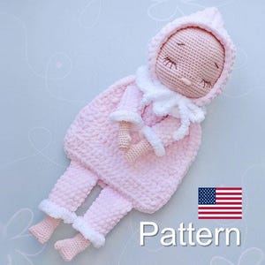 Könnte beinhalten: Eine rosa gehäkelte Puppe, die einen weißen Schlafsack mit Kapuze trägt. Die Puppe hat geschlossene Augen und ein gesticktes Lächeln. Die Puppe liegt auf einer hellblauen Oberfläche. Der Text "Pattern" ist unter der Puppe sichtbar.