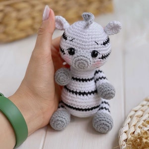 May include: A small, handmade crochet zebra toy featuring a white body with black horizontal stripes, a gray snout, gray ears, and gray paws. The plush figure has large black safety eyes, a small tuft of gray hair on top, and subtle pink blush.