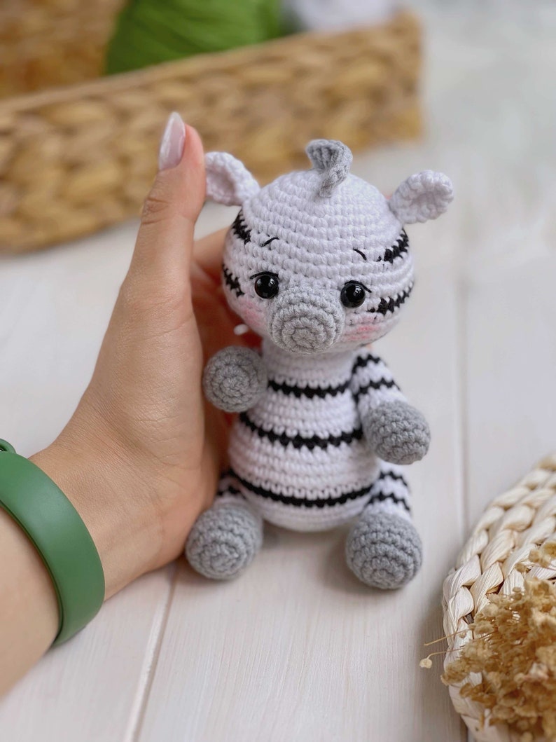 May include: A small, handmade crochet zebra toy featuring a white body with black horizontal stripes, a gray snout, gray ears, and gray paws. The plush figure has large black safety eyes, a small tuft of gray hair on top, and subtle pink blush.