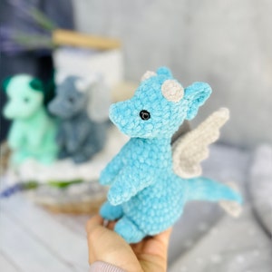 May include: A light blue crocheted dragon with white horns and wings. The plush toy has black eyes and is held in a person's hand. Other similar dragons in green and gray are in the background.