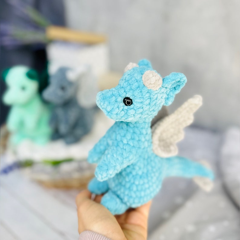 May include: A light blue crocheted dragon with white horns and wings. The plush toy has black eyes and is held in a person's hand. Other similar dragons in green and gray are in the background.